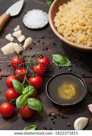 Raw maccheroni elbows pasta in glass bowl with parmesan cheese and tomatoes, oil and garlic with basil on wooden background.Macro