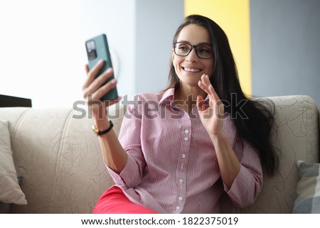 Smiling woman in glasses sits on couch, holds smartphone and waves to screen. Online dating on the internet concept Royalty-Free Stock Photo #1822375019