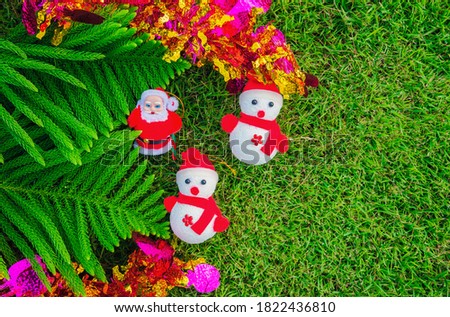 Santa Claus and Snowman on a green lawn with a Christmas tree as the background. Two snowmen paired with Santa Claus on the green grass and decorated with pine leaves.