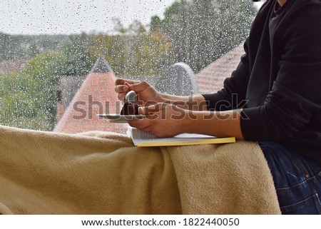 A man in his mid-twenties sits on a windowsill, in the background raindrops can be seen on the window pane - the man is reading a book while drinking a coffee - anonymous picture without head and face