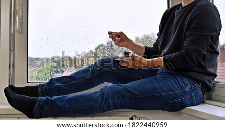 A man in his mid-twenties sits on a windowsill, in the background raindrops can be seen on the pane - the man is eating homemade chocolate cake - anonymous picture without head and face