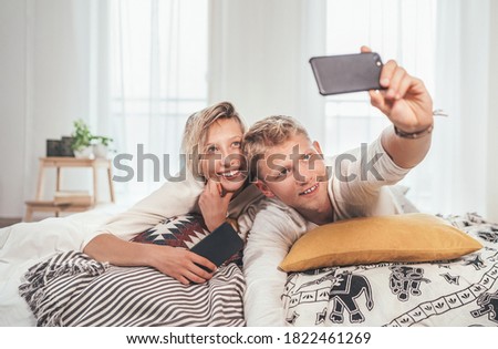 Cheerful young adults сouple in pajamas taking a selfie photo using a modern smartphone as they lazy relaxing lying in a cozy bed in the bedroom. 
 