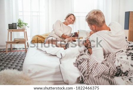 Laughing blonde hair young adults couple in pajamas lazy relaxing lying in a cozy bed in the bedroom and having a peacefully carefree chatting. Couples relations concept image.