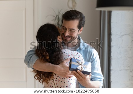 Happy young Caucasian man hug woman congratulate with anniversary present wrapped gift box. Smiling loving husband embrace wife greeting with birthday, make surprise for beloved. Relationship concept.