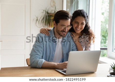 Happy Caucasian young couple look at laptop screen browse surf wireless internet on gadget. Smiling millennial man and woman shopping paying on internet on computer at home. Technology concept.