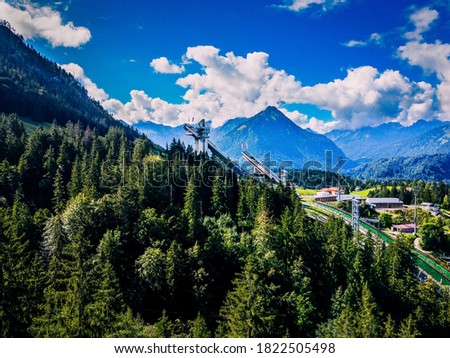 A beautiful landscape picture featuring a forrest, mountains and a ski jumping arena in the background.