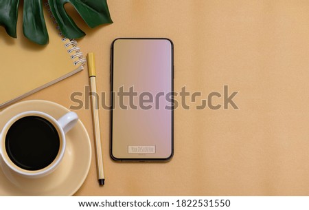 Mockup blank screen smartphone with cup of coffee, notebook and green leaf on yellow background. Copy space.