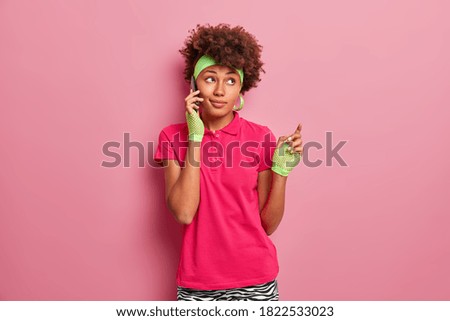 Pretty thoughtful Afro American woman with dark skin speaks on mobile phone spends free time indoors calls to friend dressed in active wear poses against pink background looks away pensively
