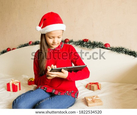 Christmas online shopping with tablet computer. Young girl wearing red sweater and Santa hat buying presents, prepare to new year, on white background near tinsels. Winter holidays sales