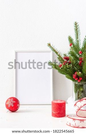 Christmas poster mock up with vertical frame, fir tree branches and hawthorn twigs in a vase and red balls with candles on white wall background, new year and christmas concept