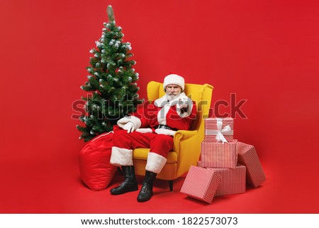 Displeased Santa Claus man in Christmas hat suit sit in armchair with fir tree presents gifts point index finger on camera isolated on red background. Happy New Year celebration merry holiday concept