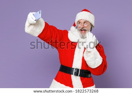 Excited Santa Claus man in Christmas hat red coat gloves glasses doing selfie shot on mobile phone greeting with hand isolated on violet background. Happy New Year celebration merry holiday concept