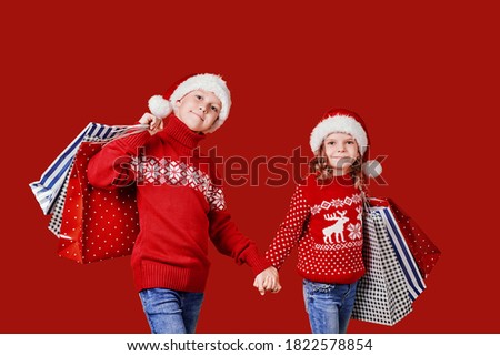 Cute children in red Santa hats, sweaters holding shopping bags on red background.