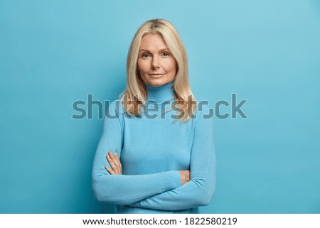 Self confident serious calm middle aged experienced blonde woman stands with arms folded dressed in casual turtleneck shows professional vibe stands in assertive pose against blue background.
