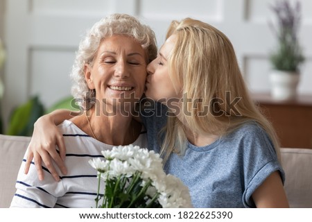 I love you so much, my dear! Affectionate millennial daughter greeting happy senior elderly mom with Mothers Day, happy grandma receiving tender kiss and flowers with closed eyes from grown grandkid