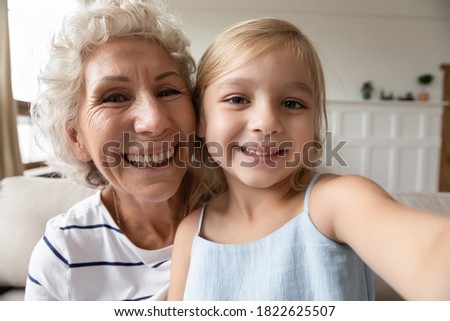 Say hi, granny! Cute little girl sitting on laps of elderly grandmother making self portrait or videocall, recording vlog, smiling preschool kid ward and older nanny looking at camera shooting video