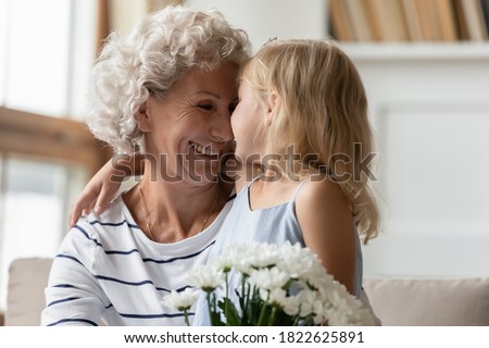 My little princess. Loving attentive little granddaughter congratulating happy mature hoary granny with birthday or Women Day cuddling her, tender looking in eyes and presenting bouquet of flowers