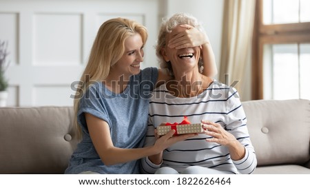 Guess what? Laughing grown up daughter congratulates happy mature mother covering her eyes and giving birthday gift, excited senior grandma receiving surprise on Mothers Day from adult granddaughter