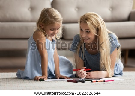 Creative activity. Happy smiling young mother spending leisure time with little kid daughter lying on heated floor in living room drawing funny pictures with bright colorful marker pens in paper album