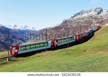 Tourists riding a Glacier Express on a brisk fall day & enjoying the view thru panoramic windows of grassy meadows on the hillside & snowy mountains under blue clear sky in Andermatt, Uri, Switzerland Royalty-Free Stock Photo #1822638128