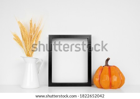 Mock up black frame with fall wheat and pumpkin decor on a white shelf. Autumn concept. Portrait frame against a white wall.