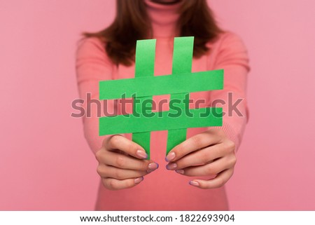 Closeup brunette woman blogger in pink sweater showing green hashtag symbol to camera recommending viral social media topic. Indoor studio shot isolated on pink background