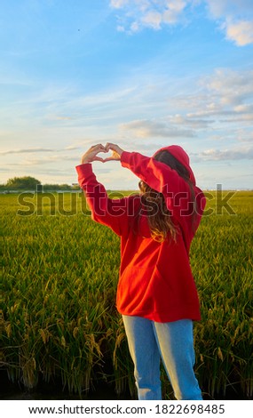 A young girl in red jacket and hood, making a heart with her hands in a rice field at sunset. Concept love, happiness, harmony, youth, color contrast.