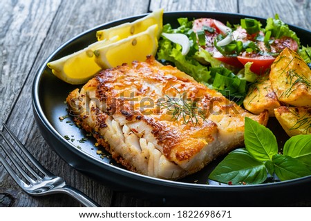 Fish dish - fried cod fillet with potatoes and vegetable salad on wooden table 