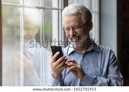 Happy senior Caucasian man in glasses look at cellphone screen browse wireless internet. Smiling modern mature 70s grandfather text or message on smartphone. Elderly use cell technologies at home.
