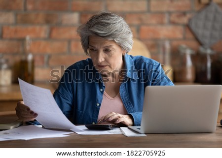 Serious mature Caucasian 60s woman sit at table calculate household finances paying bills on laptop online. Focused senior grey-haired grandmother manage family home budget, read paper bank notice Royalty-Free Stock Photo #1822705925
