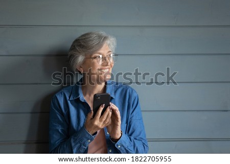 Happy modern senior 60s woman in glasses isolated on grey wooden wall background use smartphone look in distance. Smiling mature female text message on cellphone dreaming or thinking at home.