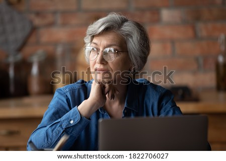 Pensive mature grey-haired Caucasian woman in glasses distracted from computer work thinking or dreaming. Dreamy elderly 60s female in spectacles at home look in distance make plans or decisions. Royalty-Free Stock Photo #1822706027