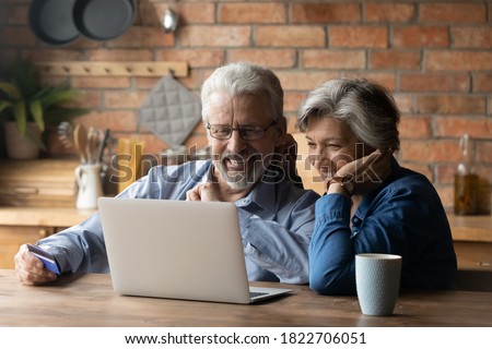 Smiling mature 70s couple sit relax at table at home kitchen have fun shopping online on computer together. Smart modern happy senior man and woman look at laptop screen, pay with credit card o web. Royalty-Free Stock Photo #1822706051