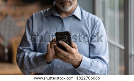 Crop close up of mature Caucasian grandfather hold modern smartphone gadget browse wireless internet. Senior man look at cellphone screen, read news online on device. Elderly and technology concept. Royalty-Free Stock Photo #1822706066