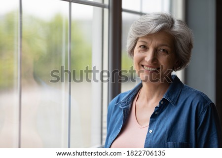 Portrait of smiling mature Caucasian 60s woman stand near window in own house show optimism at retirement. Profile picture of happy elderly female feel positive overjoyed, enjoy good days at home.