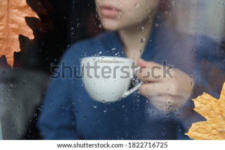 Woman drinking hot tea or coffee and looking through window with raindrops . Window background . Autumn leaves . White cup of tea or coffee .