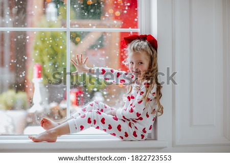Young pretty girl with long blond curly hair and red Christmas bow in white winter pyjamas in red hearts posing on the big window with snow background.