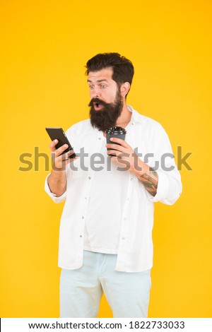 guy with beard and moustache hold mobile phone. communication. morning routine. surprised bearded man has incoming phone call while drink coffee. brutal hipster drinking takeaway beverage from cup.