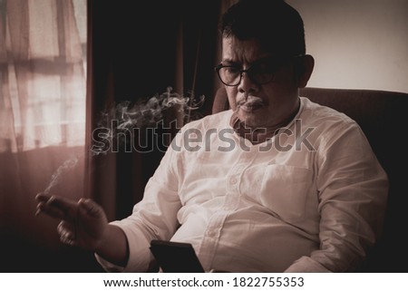 Man like a businessman or leader, is using a smartphone while sitting and smoking in a modern room against  the sunlight behind the window as background.