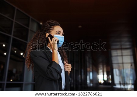 Portrait of a young attractive woman talking business on the mobile phone wearing a medical sterile mask outside the office building. Leading business during Covid-19 pandemic concept. Royalty-Free Stock Photo #1822766021