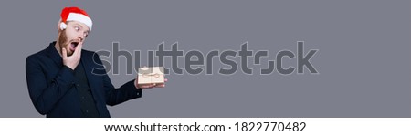 Bearded caucasian man with a santa hat is gesturing surprisement while holding a box with present on a gray studio wall with free space