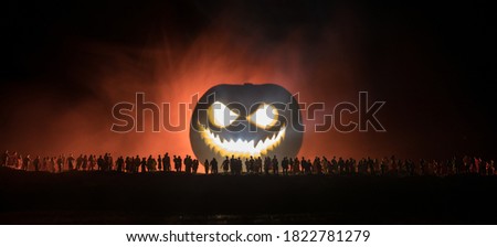 Halloween concept. Blurred silhouette of giant Jack-o-lantern pumpkin with scary smiling face behind crowd at night. People looks at a big pumpkin at night. Selective focus.