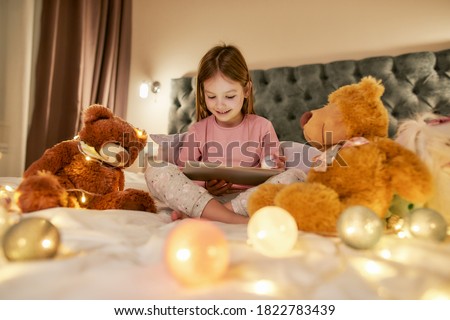 A cute small girl wearing pyjamas sitting alone cross-legged on a huge bed barefoot, watching cartoons on a tablet and smiling before going to sleep in a big well-lighted bedroom