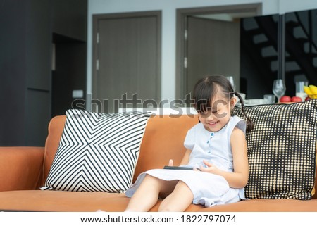 Asian cute child using a smartphone and smiling for watching video or play game while sitting on sofa in living room at home