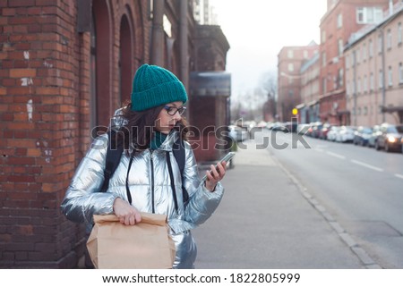 courier or volunteer carries food in a paper bag. A young woman in a warm jacket holds a parcel and uses a smartphone