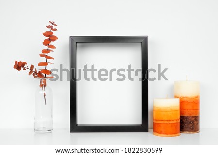 Mock up black frame with fall branches and candle decor on a white shelf. Autumn concept. Portrait frame against a white wall.
