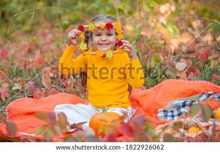 Little cute girl in an orange T-shirt holds a wooden frame and looks through it, the frame is decorated with autumn leaves and pumpkins cut from paper, autumn composition, children's creativity.
