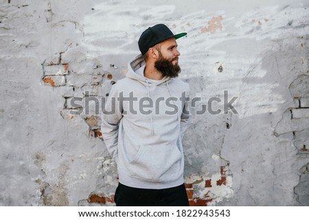 City portrait of handsome hipster guy with beard wearing gray blank hoodie and baseball cap with space for your logo or design. Mockup for print