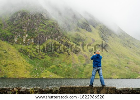 Bald tourist taking picture on his smart phone of a beautiful scenery. Connemara region, Ireland. Lake and mountain covered with cloud in the background. Man dressed in blue hoodie and jeans.