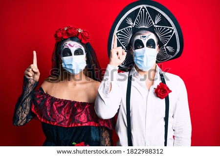 Couple wearing day of the dead costume wearing medical mask pointing finger up with successful idea. exited and happy. number one. 